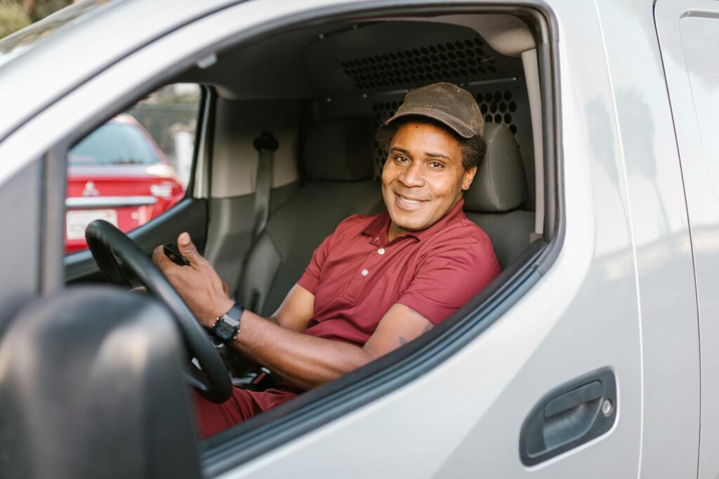 A smiling delivery driver in uniform seated inside his van, holding a smartphone.